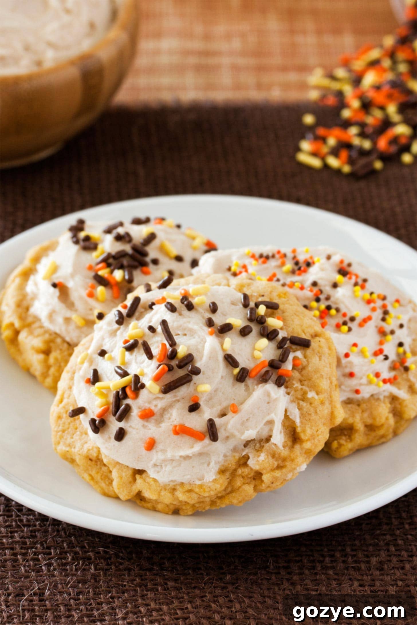 Autumn Spice Delights 24 Close-up of three soft pumpkin sugar cookies with cinnamon buttercream and festive sprinkles on a white plate