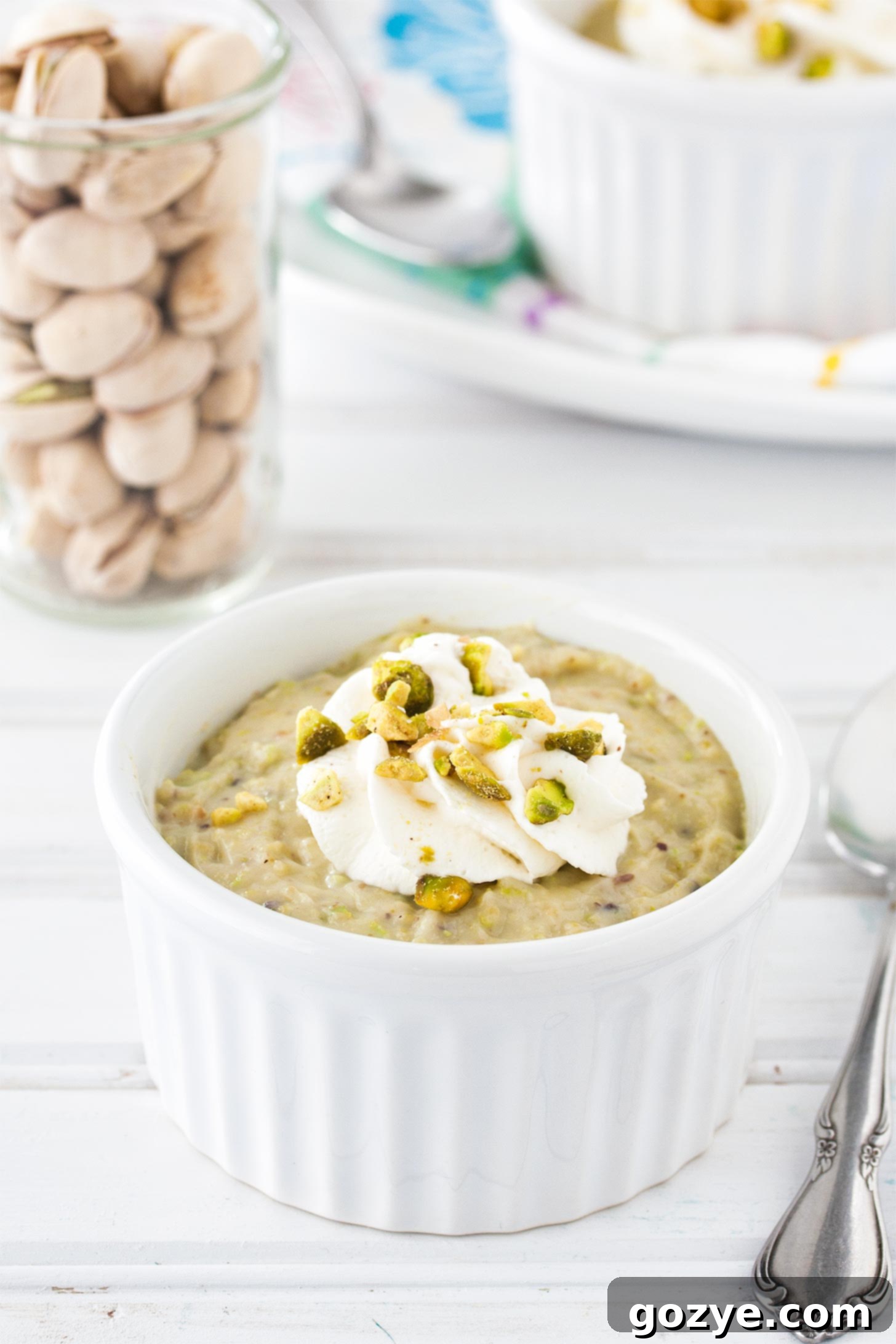 A serving of homemade pistachio pudding in a white ramekin, with a spoon gently placed on the side, inviting a taste. The pudding is a subtle, natural green color, showcasing its authentic ingredients.