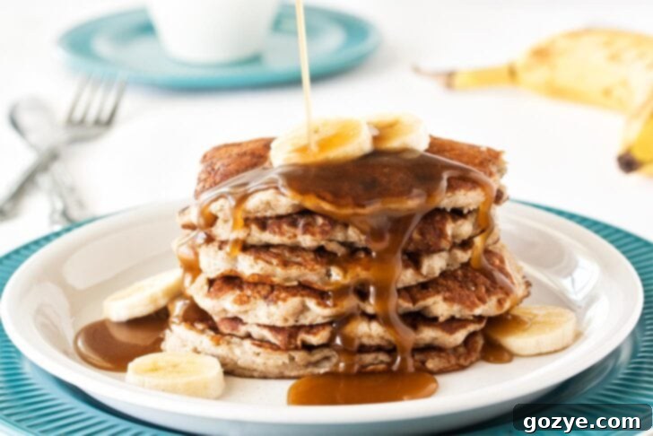 Dairy-Free Banana Coconut Milk Pancakes 4 A hand pouring maple syrup over a stack of fluffy coconut milk pancakes with banana slices, emphasizing the delicious topping options.