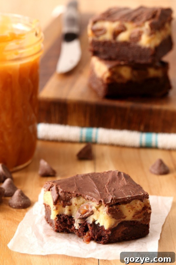 A perfectly cut slice of Caramel Chocolate Chip Cookie Dough Brownies displayed on a white surface, highlighting its three layers.