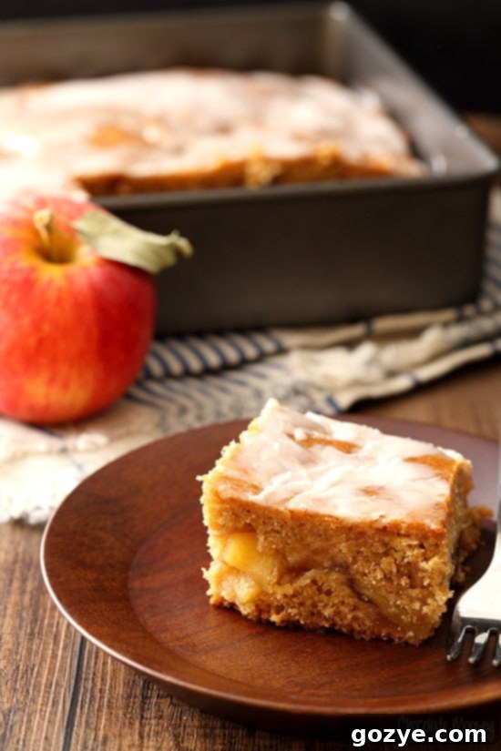 A delectable slice of Apple Fritter Cake resting on a rustic wooden plate, revealing layers of moist cake and rich apple pie filling, drizzled with sweet glaze.