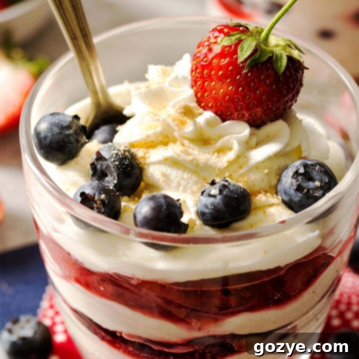Close up of two No Bake Cheesecake Cups, with a spoon in one, showing layers of creamy filling, graham cracker crust, strawberries, and blueberries.