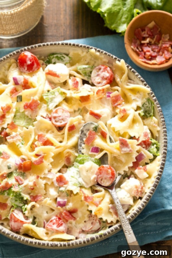 Large bowl of BLT Pasta Salad with a serving spoon, garnished with fresh herbs and visible bacon, tomatoes, and mozzarella