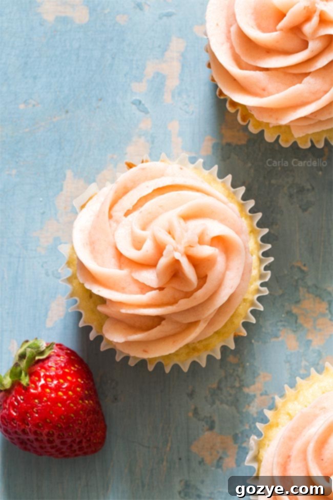 Close-up of a vanilla cupcake generously frosted with pink strawberry buttercream, garnished with a fresh strawberry slice.