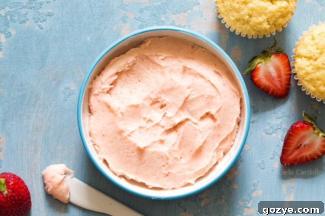 A large bowl of smooth, pale pink strawberry buttercream frosting, topped with a fresh, vibrant red strawberry.