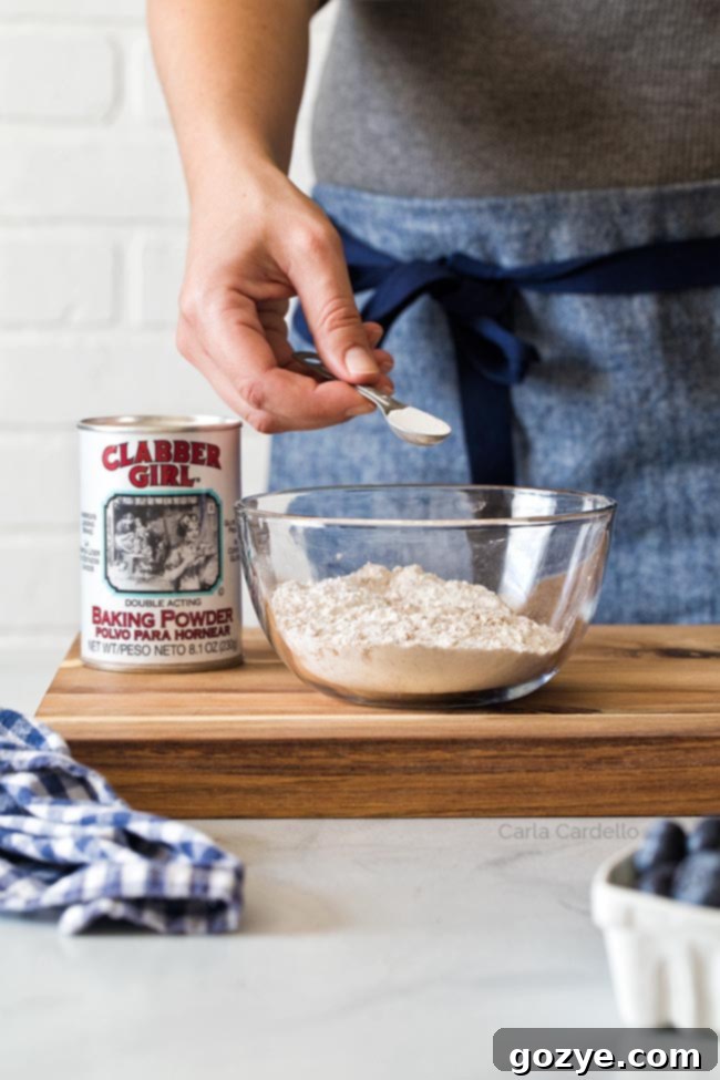 A hand adding half a teaspoon of baking powder into a bowl of all-purpose flour, alongside other dry ingredients
