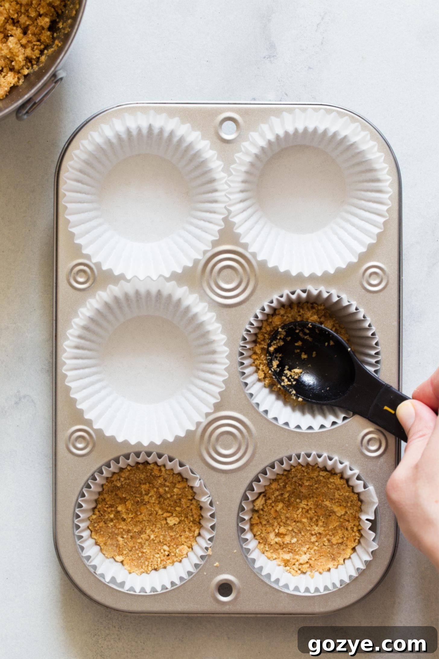 Quick No-Bake Cheesecake Bites 6 Hands pressing graham cracker crumbs into paper liners in a muffin pan to form mini crusts