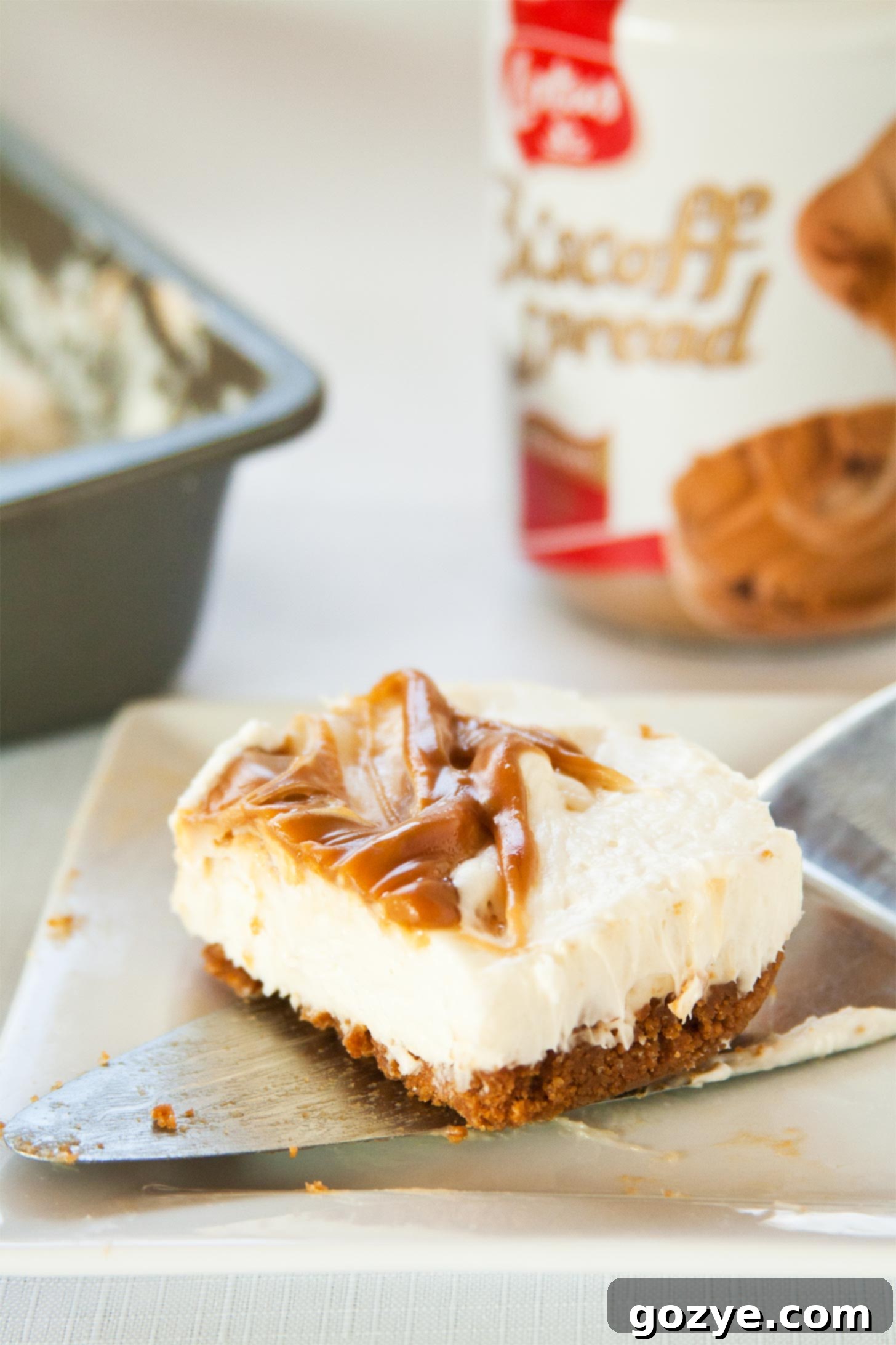 No Bake Cookie Butter Cheesecake being scooped from an 8x8 pan onto a serving plate, highlighting a clean slice