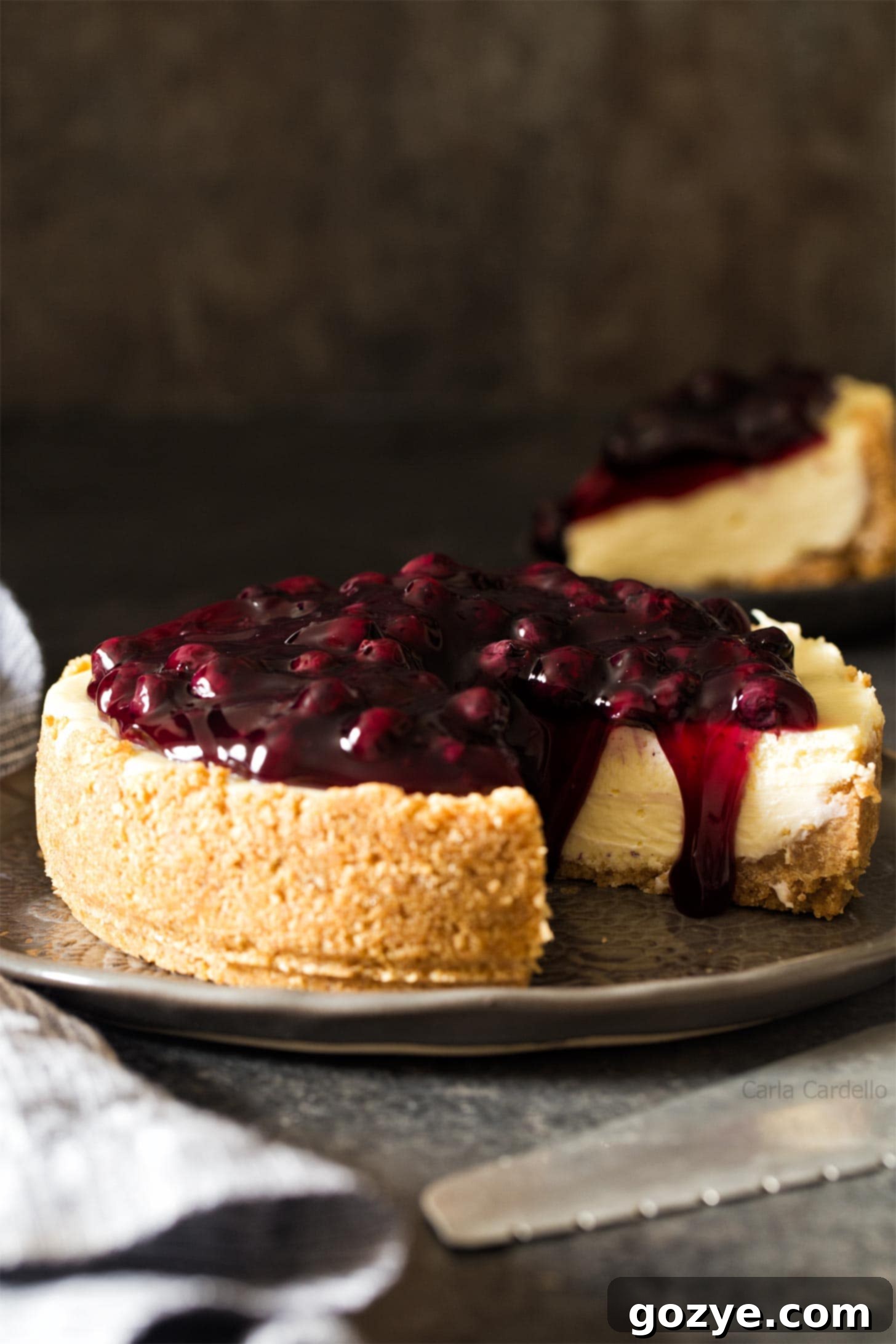 A whole 7 inch cheesecake adorned with blueberry pie filling on a stylish gray plate, ready to be served.