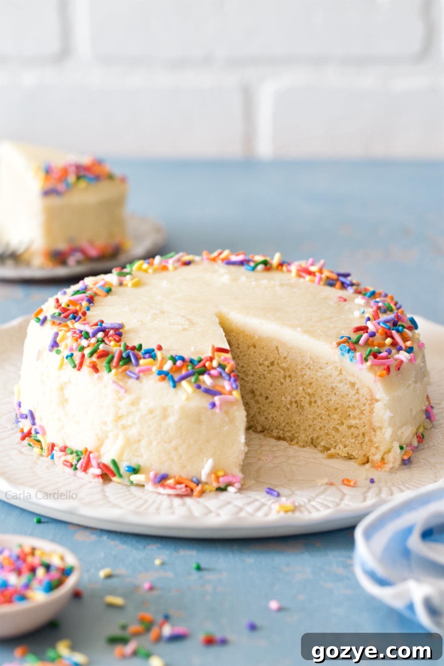 A small vanilla cake beautifully frosted, resting on a white platter on a blue table, highlighting its elegant simplicity.