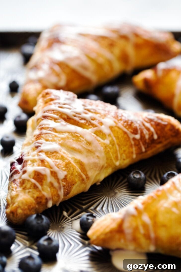 Close up of a freshly baked blueberry turnover on a baking tray, showing its golden-brown, flaky crust and a hint of the juicy blueberry filling peeking through.