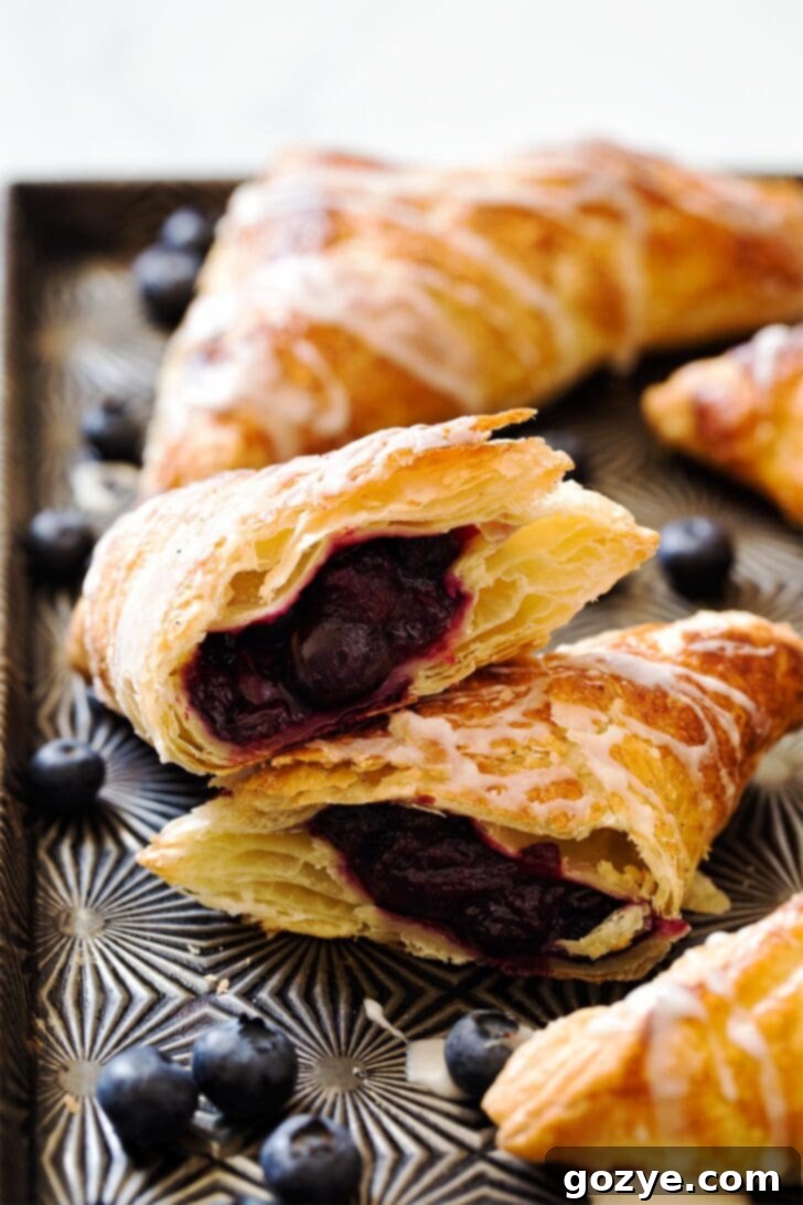 Two puff pastry blueberry turnovers, one cut in half to reveal the rich, purple blueberry pie filling inside, resting on a rustic wooden surface.