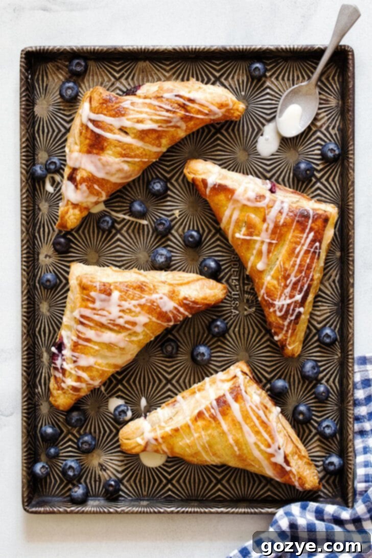 An overhead shot of four perfectly baked blueberry turnovers arranged on a cooling rack, glistening with a fresh vanilla glaze drizzle.