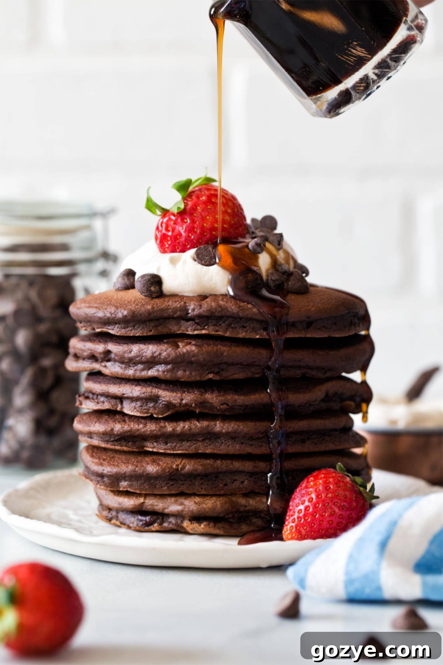 Pouring rich maple syrup over a tall stack of fluffy double chocolate pancakes with whipped cream and fresh berries