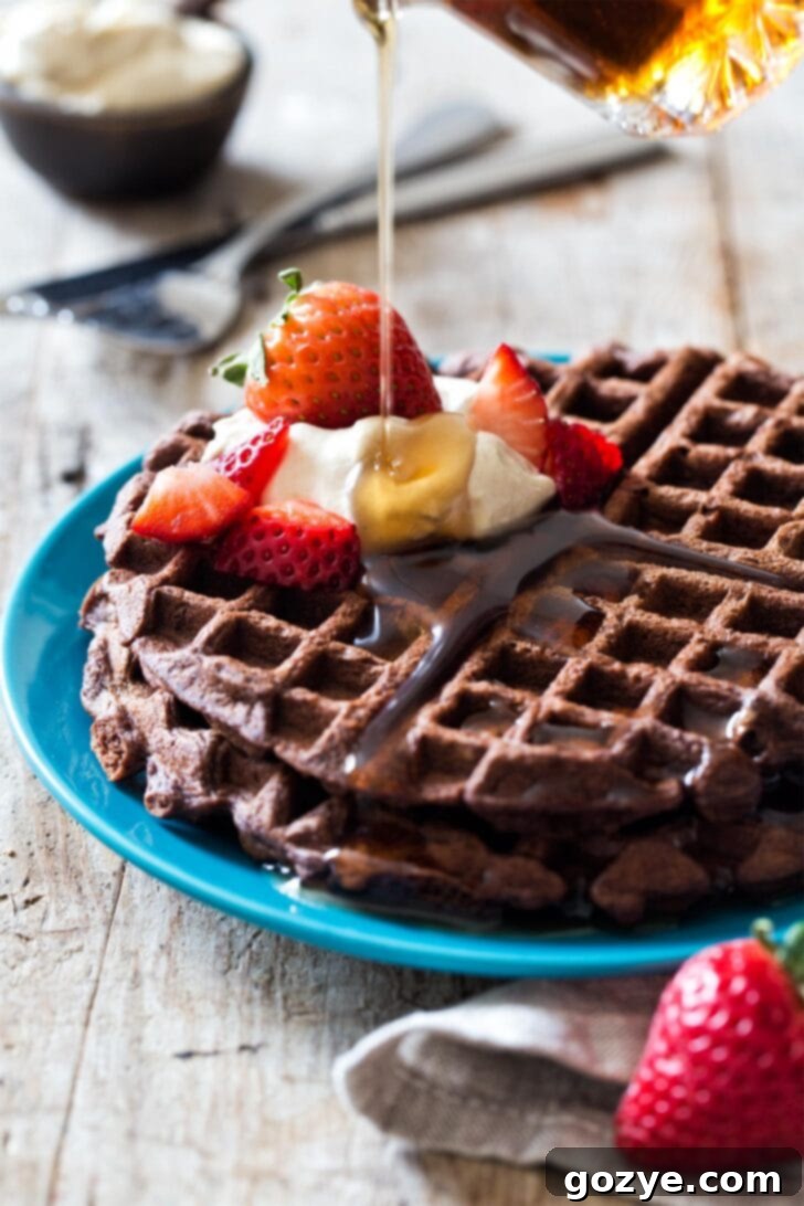 Pouring maple syrup generously over freshly made, crispy chocolate waffles on a white plate, highlighting their texture.