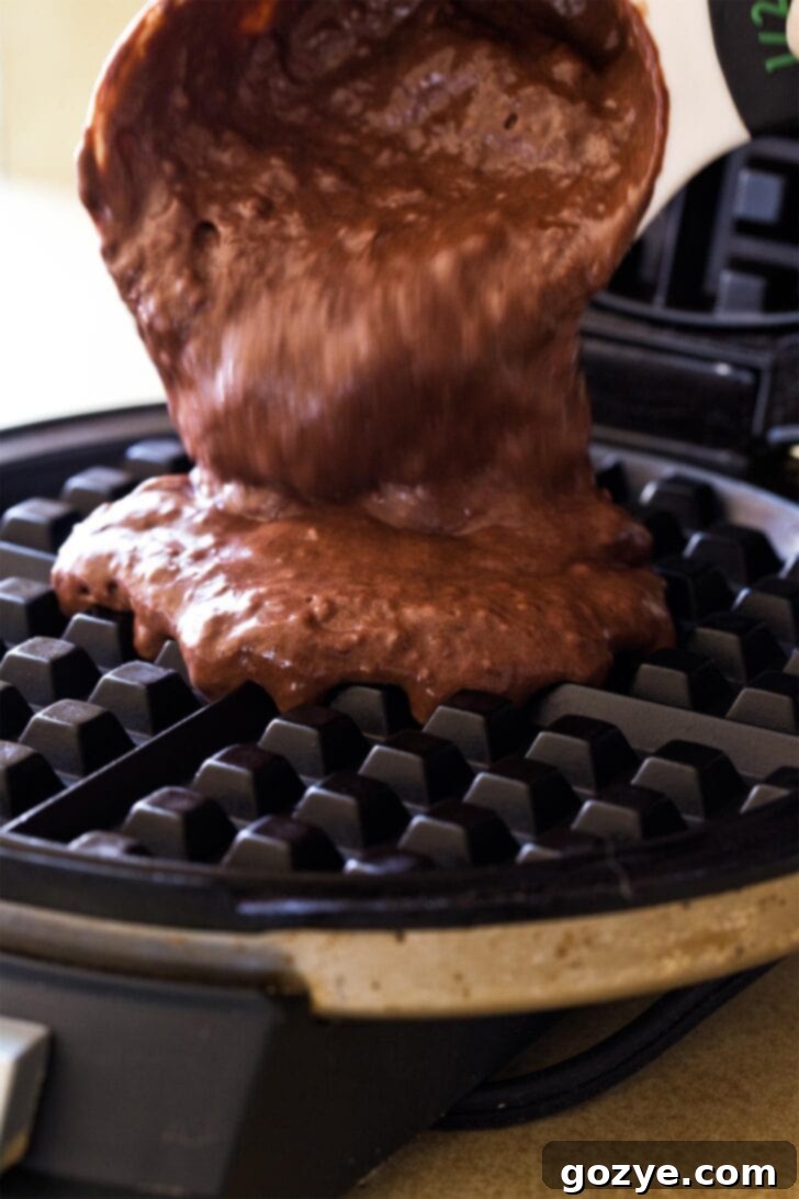 A ladle dispensing thick chocolate waffle batter evenly onto the hot plates of a preheated waffle maker.