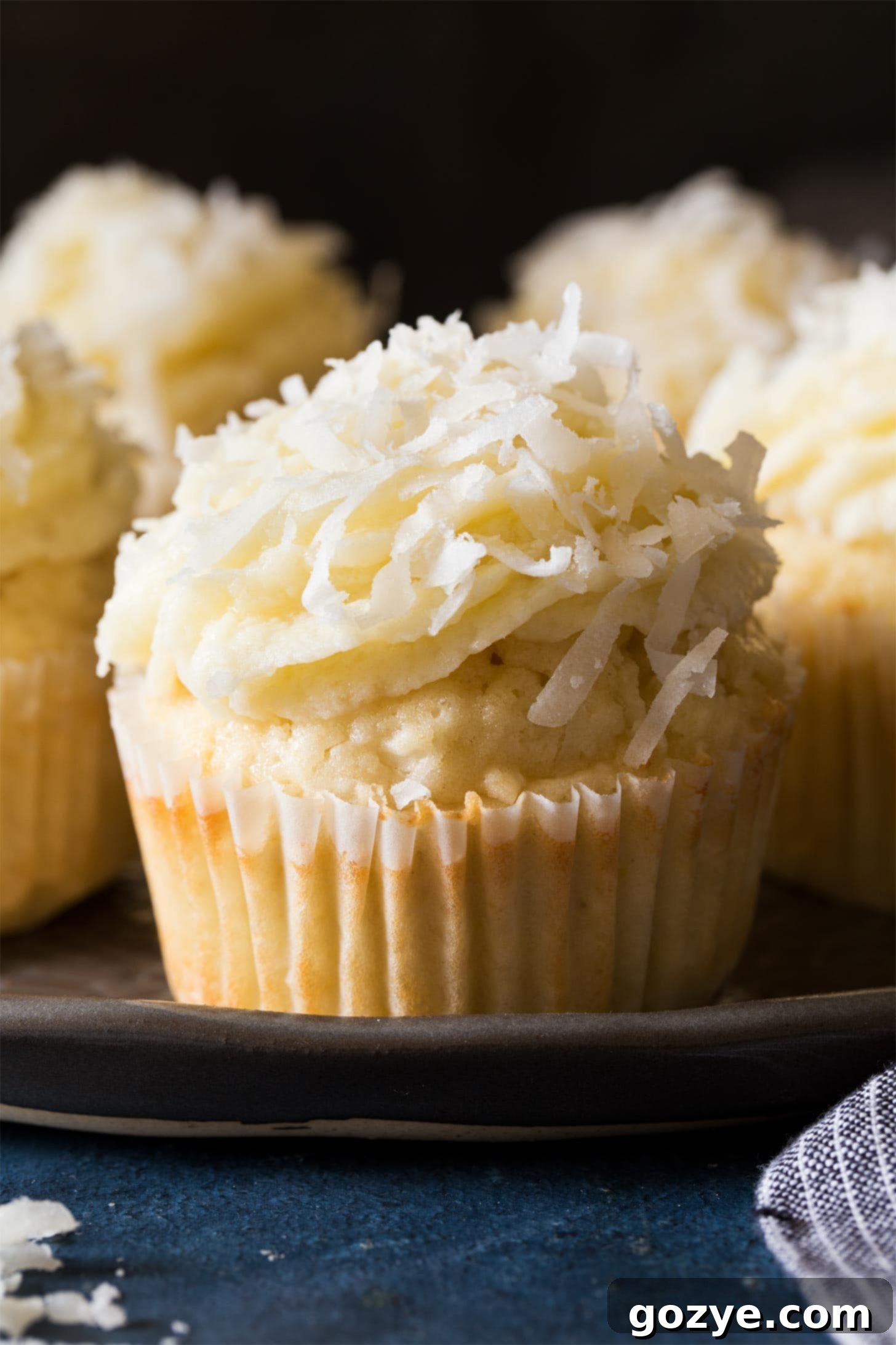 Close up of a frosted coconut cupcake on a white plate, showing the fluffy texture and sprinkled coconut topping.