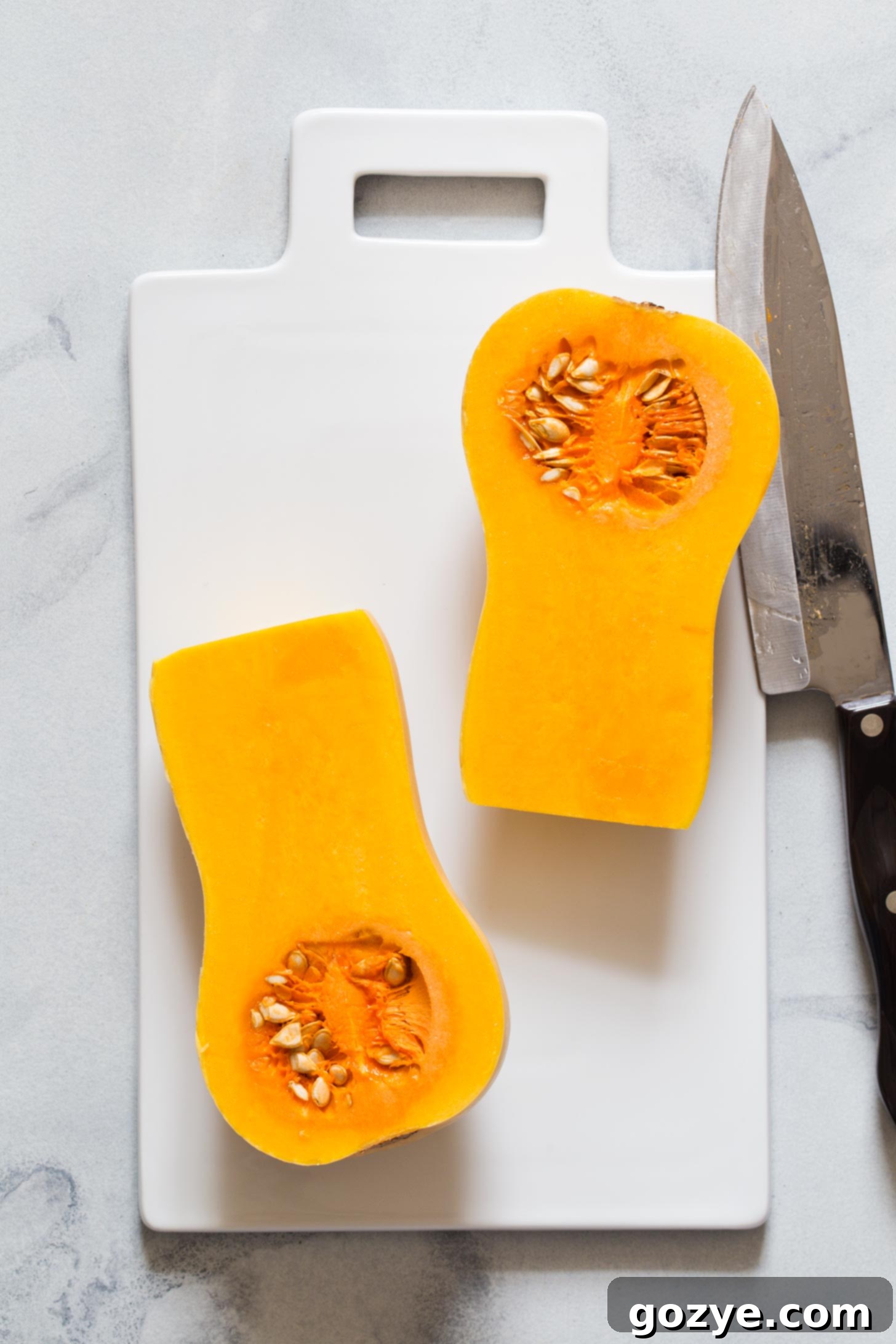 A butternut squash being carefully cut in half lengthwise on a wooden cutting board for roasting